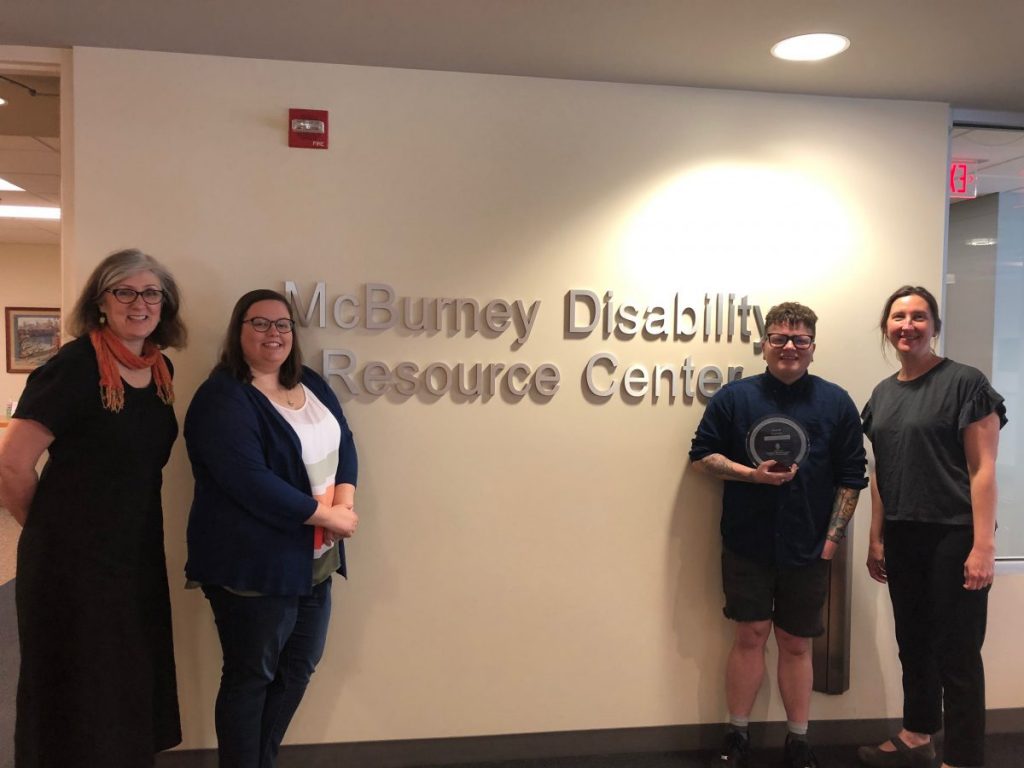 Photo of Peggy Weaver, Kate Doran, Caelyn Randall, and Amy Free standing in front of the McBurney Disability Resource Center wall sign