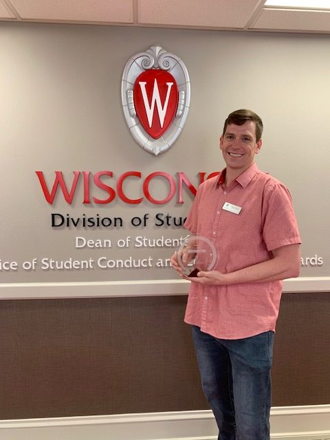 Photo of Tony Utrie holding award standing in front of Dean of Students wall logo