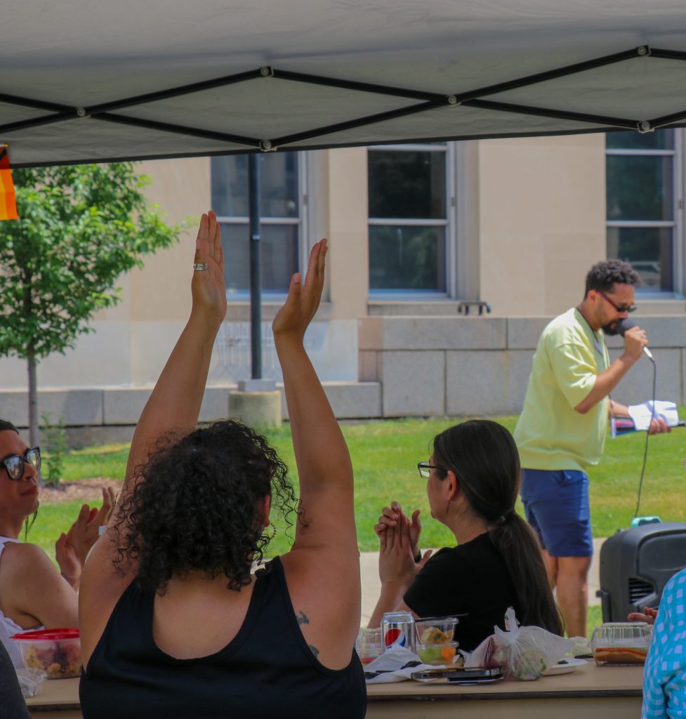 Director of the Gender and Sexuality Campus Center, Warren Scherer (Center), speaks in front of the attendees of the Pride Picnic. Attendees cheer and clap their hands.