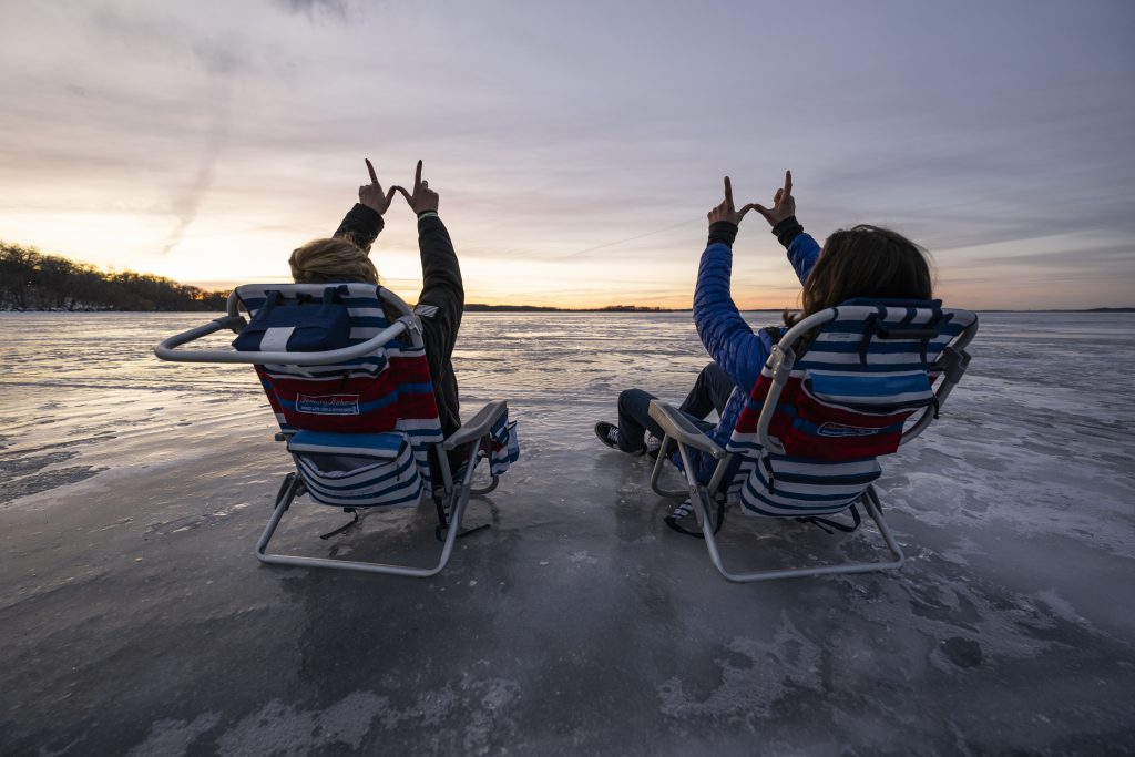 Two students throw W hand signs after setting up a pair of lawn chairs on frozen and ice-covered Lake Mendota.