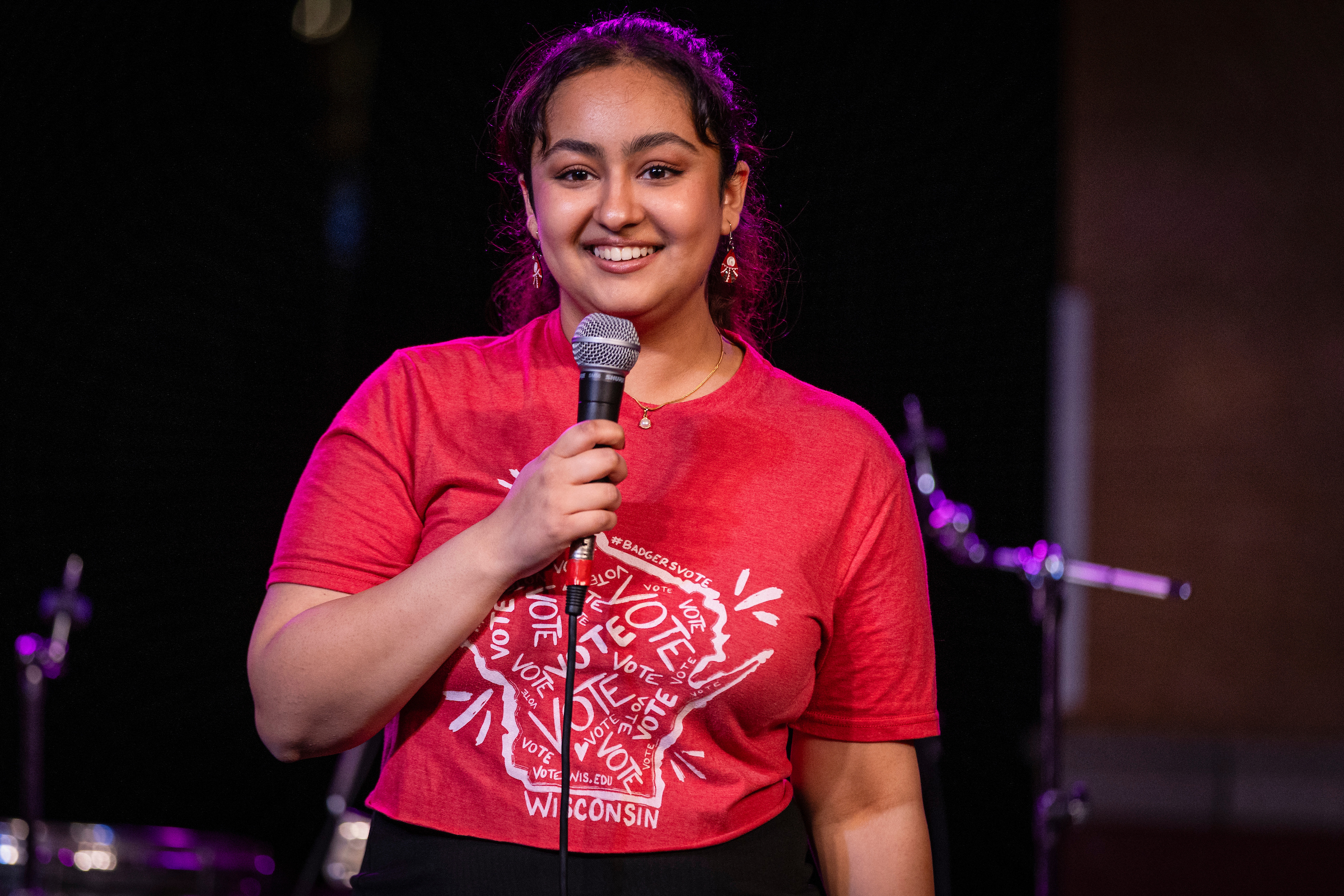 Amanjot Kaur stands with a microphone, wearing a red shirt that says "Vote" with an outline of the state of Wisconsin" while attending Voterpalooza 2024.