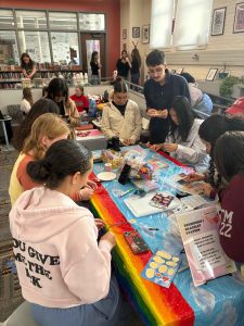 Students gather to celebrate National Coming Out Day. Students are pictured around a table making friendship bracelets. 