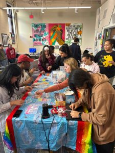 Students gather to celebrate National Coming Out Day. Students are pictured around a table making friendship bracelets. 