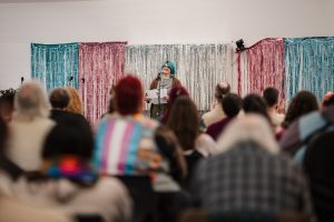 Camren Livermore, a white performer with blue hair, glasses, and sunflower earrings, stands at a microphone smiling at the crowd. He is in front of a backdrop of blue, pink, and silver metallic fringe backdrop. Several rows of audience members are blurry in the foreground of the picture, including one wearing a trans flag on the back of their jacket.