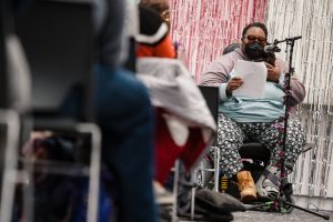 T. S. Banks, a fat, Black, trans performer with long locs, sits in his power chair and reads off a sheet of paper at a microphone wrapped in purple flowers. He is wearing a black KN94 mask and tinted glasses. He is in front of a backdrop of metallic pink and silver fringe. Rows of audience members are visible on the left side.