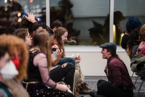 Audience members sit in rows. The wall behind them is reflective windows at night. Zoe Loschke, a white attendee with a long braid and wearing a grey newsboy cap, sits on the floor talking with seated attendees. Everyone is smiling.