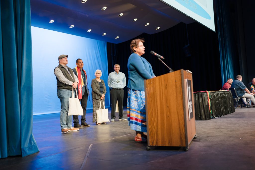 APiO sister, Adrienne Thunder addresses the audience at the podium. Behind her are her fellow All City founders, standing together.