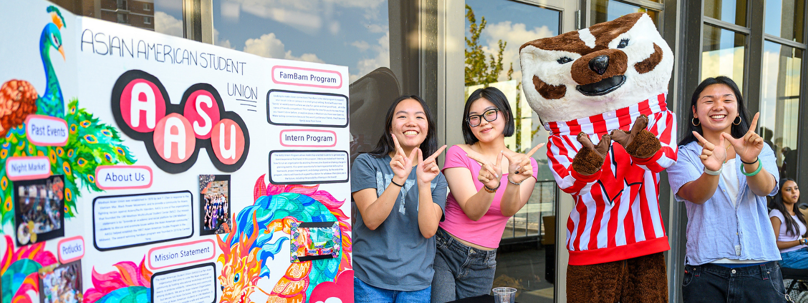 A group of students show a 'W' with Bucky in front of their table display about the Asian American Student Union (AASU).