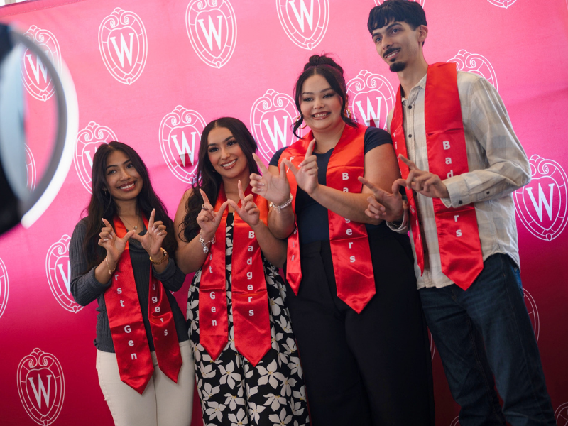 Four students pose for a photo in the photo booth, they have stoles on that read 'First Gen Badger'